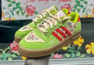 A pair of vibrant green and beige sneakers with floral embroidery and red accents, featuring the Adidas logo and brown soles, displayed against a background of colorful floral artwork.