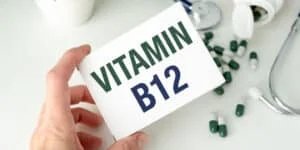 A hand holding a box labeled 'VITAMIN B12' with green text, surrounded by scattered green and white capsules and a stethoscope in the background.