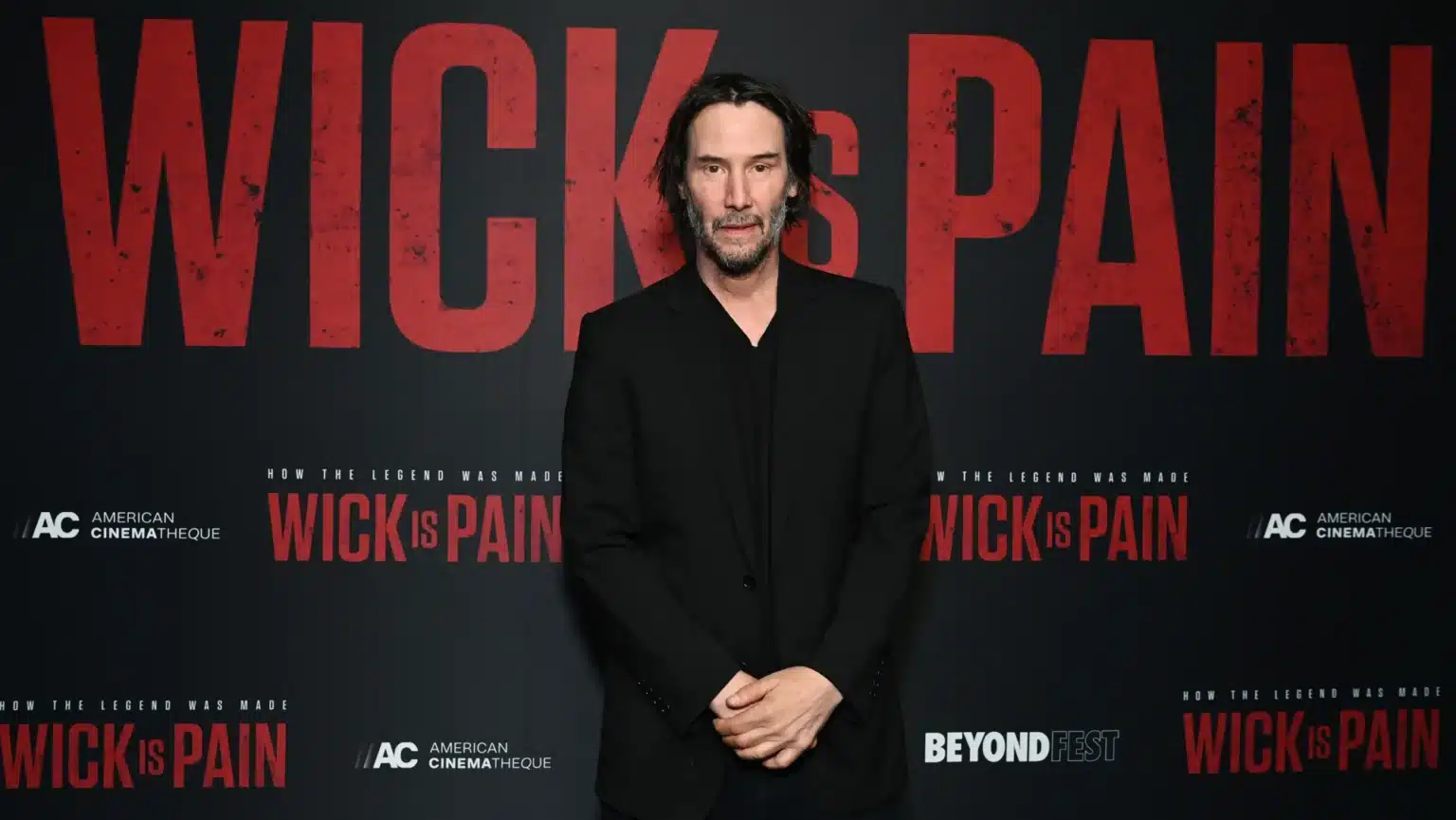 A man in a black suit stands in front of a promotional backdrop featuring bold red text that reads "WICK is PAIN". He has long, dark hair and a serious expression.