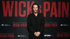 A man in a black suit stands in front of a promotional backdrop featuring bold red text that reads "WICK is PAIN". He has long, dark hair and a serious expression.