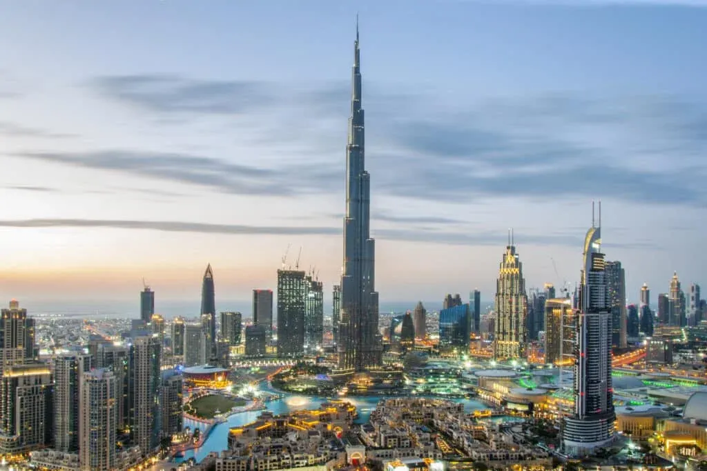 A panoramic view of Dubai's skyline at dusk, featuring the Burj Khalifa prominently in the center, surrounded by various modern skyscrapers and illuminated buildings, with waterways and greenery below.