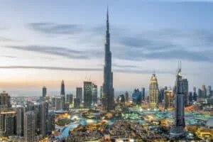 A panoramic view of Dubai's skyline at dusk, featuring the Burj Khalifa prominently in the center, surrounded by various modern skyscrapers and illuminated buildings, with waterways and greenery below.