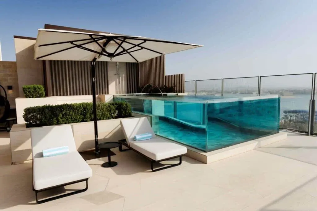 A stylish rooftop terrace featuring a clear infinity pool with an underwater view, two white lounge chairs with a turquoise towel, an umbrella providing shade, and trimmed hedges in the background, set against a clear blue sky.