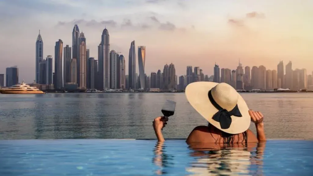 A woman in a large straw hat relaxes in an infinity pool, holding a glass of red wine, with a view of a skyline featuring tall skyscrapers and a sunset over the water.