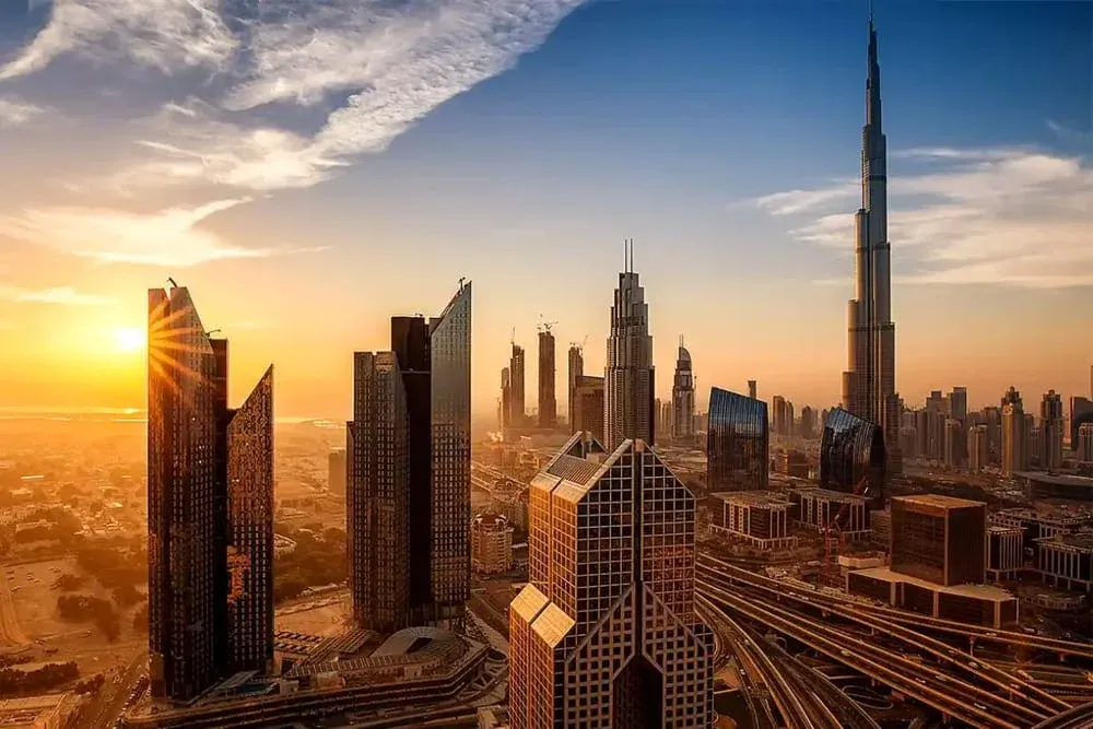 A panoramic view of a modern city skyline at sunset, featuring several skyscrapers including the Burj Khalifa, with the sun shining through the buildings, casting long shadows and highlighting their architectural details.