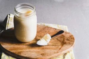 A glass mason jar containing a creamy substance, placed on a wooden cutting board, with a spoonful of the same substance next to it; a striped kitchen towel is partially visible in the background.