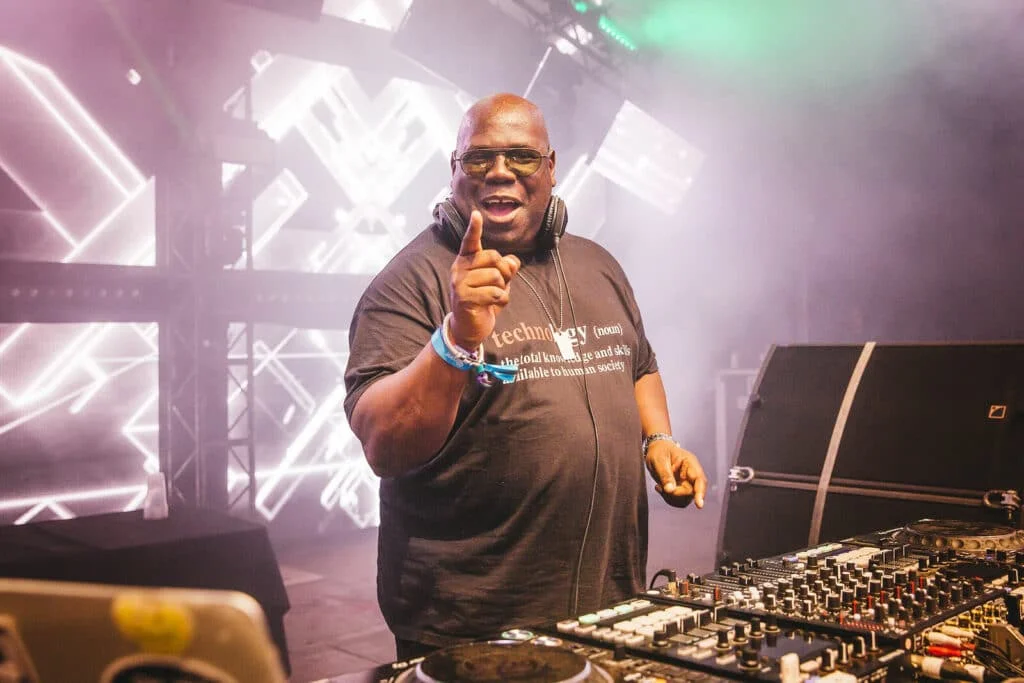 A DJ, wearing sunglasses and a black t-shirt with a definition of 'technology' printed on it, raises a finger while performing at a festival. He stands behind a DJ booth with various equipment and colorful stage lighting in a smoky atmosphere.