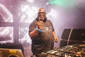 A DJ, wearing sunglasses and a black t-shirt with a definition of 'technology' printed on it, raises a finger while performing at a festival. He stands behind a DJ booth with various equipment and colorful stage lighting in a smoky atmosphere.