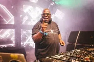 A DJ, wearing sunglasses and a black t-shirt with a definition of 'technology' printed on it, raises a finger while performing at a festival. He stands behind a DJ booth with various equipment and colorful stage lighting in a smoky atmosphere.