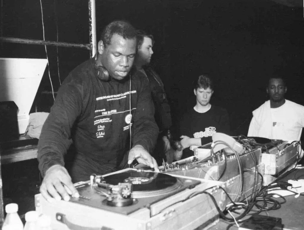 A DJ with headphones is focused on mixing records at a turntable, while three men observe in the background, each displaying a range of expressions in a dimly lit setting.