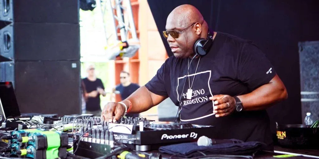 A DJ performing on stage with a turntable setup, wearing sunglasses and a black t-shirt that says 'Techno Religion', focused on mixing music with an audience visible in the background.