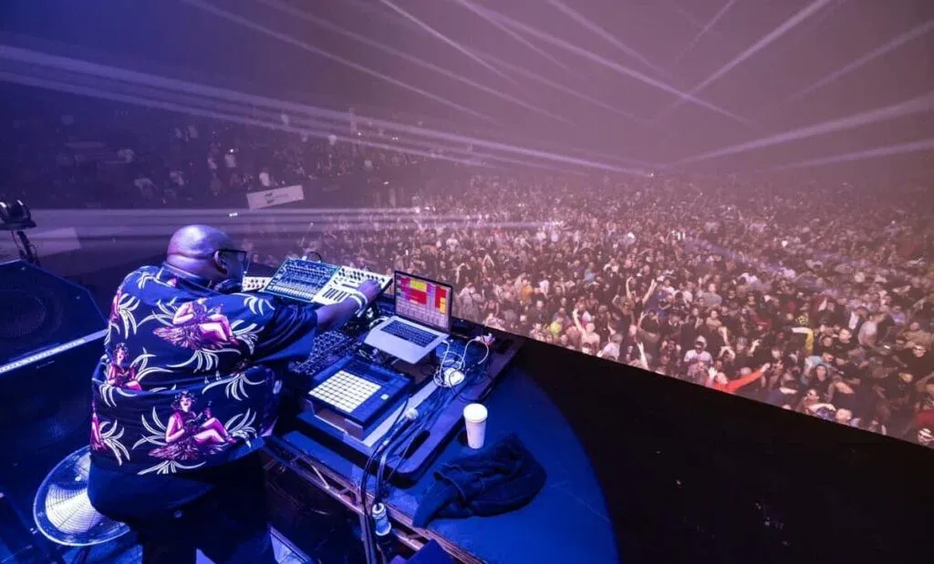 A DJ performs on stage, wearing a colorful patterned shirt, and operating electronic equipment while a large, enthusiastic crowd can be seen in the background, illuminated by stage lights.