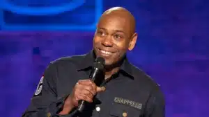 A smiling comedian stands on stage holding a microphone, wearing a dark shirt with 'CHAPELLE' embroidered on it, with a colorful background.