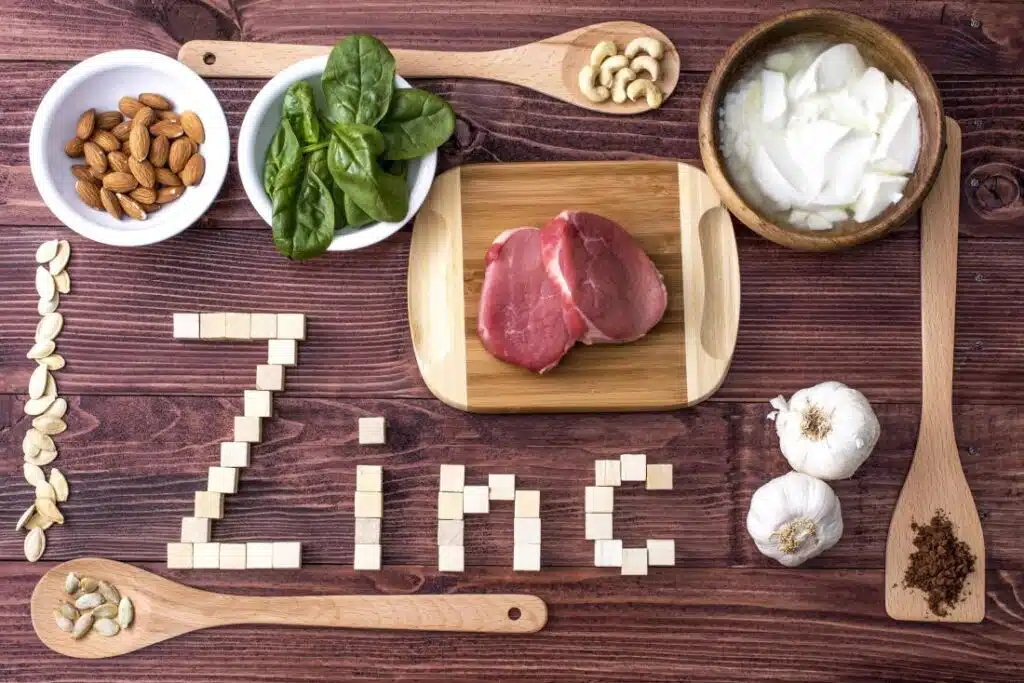 An arrangement of various foods including almonds, spinach, cashews, yogurt, garlic, and a piece of meat, with the word 'Zinc' spelled out in wooden blocks and pumpkin seeds on a wooden surface.