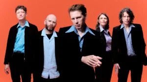 A group of five musicians posing against a vibrant orange background, wearing matching black jackets over light blue shirts, showcasing a mix of styles and expressions.