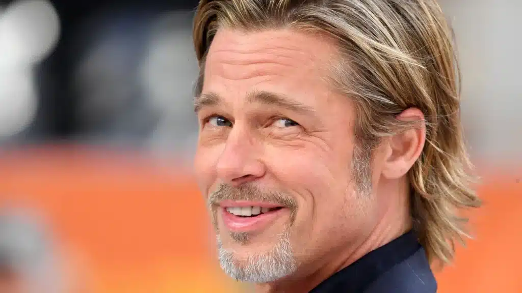 A smiling man with light brown hair and a goatee, wearing a dark blazer, poses for the camera against a blurred background with hints of orange and bokeh effects.