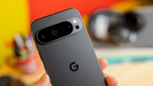 Close-up of the back of a smartphone held in a hand, featuring a prominent camera module with multiple lenses and a flash, against a blurred background of vibrant colors and various office supplies.