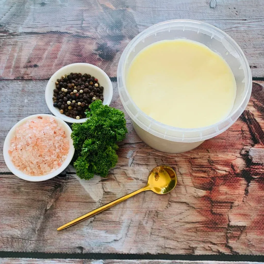 A container of cream or sauce sits on a wooden surface, surrounded by bowls of pink salt and mixed peppercorns, a sprig of fresh parsley, and a golden spoon.