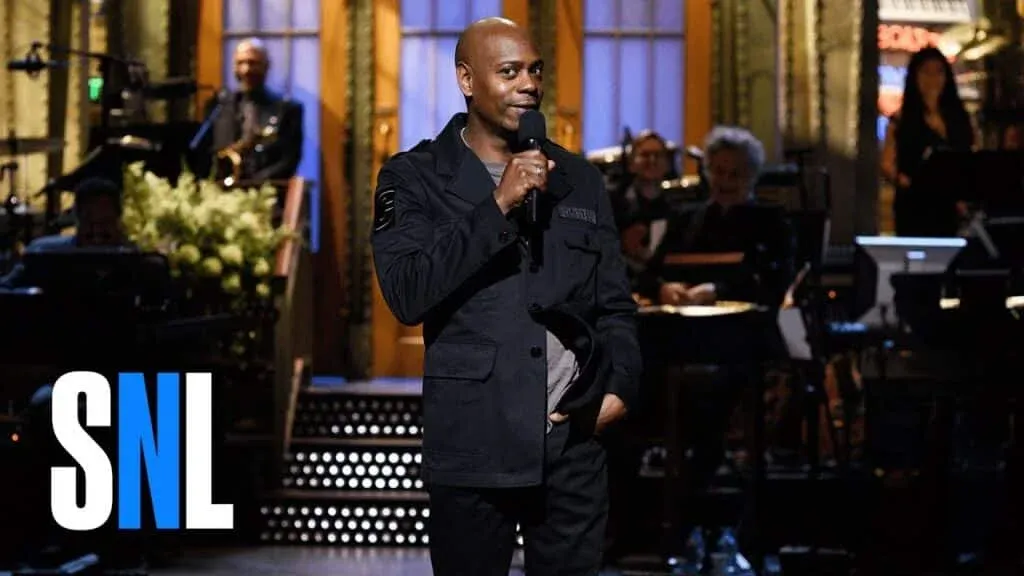 A comedian standing on stage during a live performance, wearing a dark jacket and holding a microphone, with a band and audience in the background, and 'SNL' logo in the lower left corner.