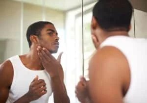 A man in a white tank top stands in front of a mirror, inspecting his face and grooming his facial hair.