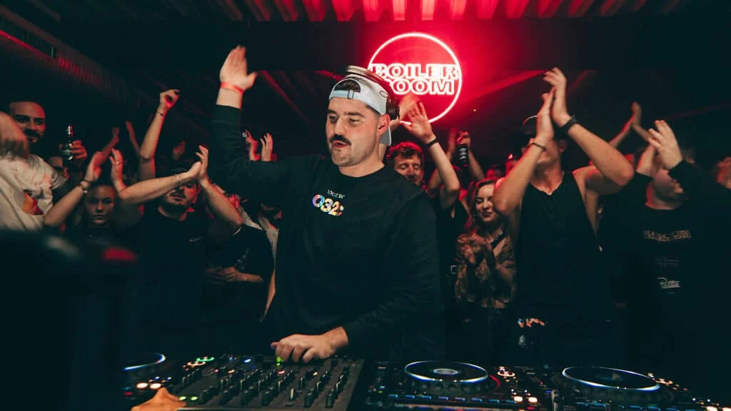 A DJ wearing a cap performs in front of a lively crowd during a music event, with many people clapping and enjoying the atmosphere, illuminated by red lighting and a 'BOILER ROOM' sign in the background.