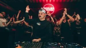 A DJ wearing a cap performs in front of a lively crowd during a music event, with many people clapping and enjoying the atmosphere, illuminated by red lighting and a 'BOILER ROOM' sign in the background.