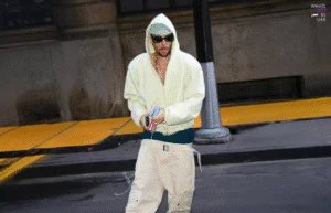 A person dressed in a light yellow hoodie and cream-colored baggy pants is walking on a city street. They are wearing sunglasses and holding a handheld device with a cord. The background features a dark pavement with yellow stripes and a tall building facade.