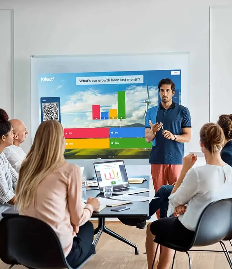 A man presenting to a group seated around a conference table, with a large display screen behind him showing a colorful bar graph and a question about growth over the last month. The audience appears engaged, with laptops and documents on the table.