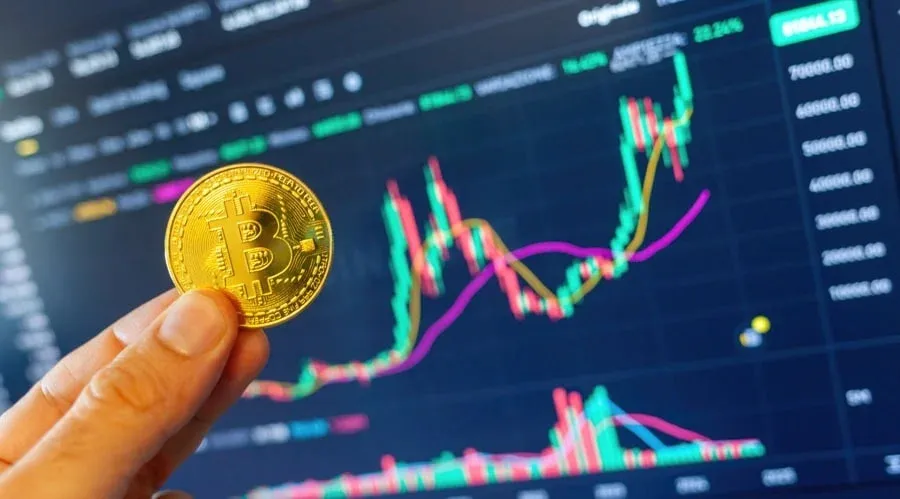 A person holding a gold Bitcoin in front of a digital cryptocurrency trading chart displaying various price movements and trends.