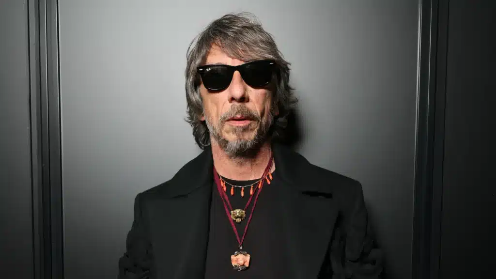 A man with shaggy gray hair and sunglasses stands against a dark background. He wears a black coat and multiple necklaces with various pendants.