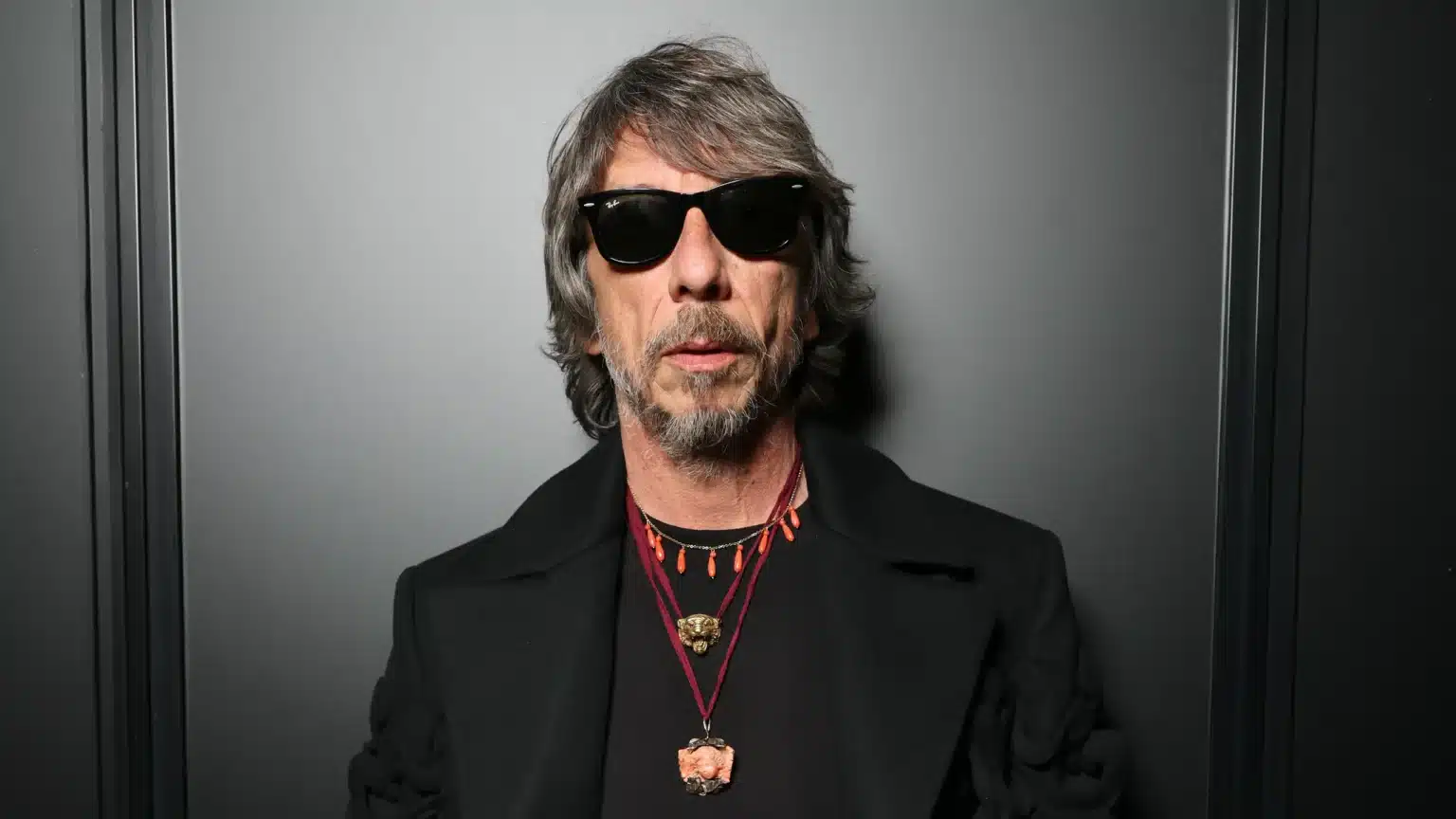 A man with shaggy gray hair and sunglasses stands against a dark background. He wears a black coat and multiple necklaces with various pendants.
