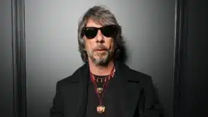 A man with shaggy gray hair and sunglasses stands against a dark background. He wears a black coat and multiple necklaces with various pendants.