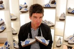 A person sitting among shelves filled with various white sneakers featuring blue stripes, holding a pair of these sneakers, with a neutral expression.