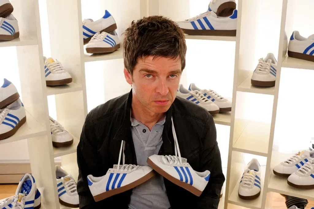 A person sitting among shelves filled with various white sneakers featuring blue stripes, holding a pair of these sneakers, with a neutral expression.