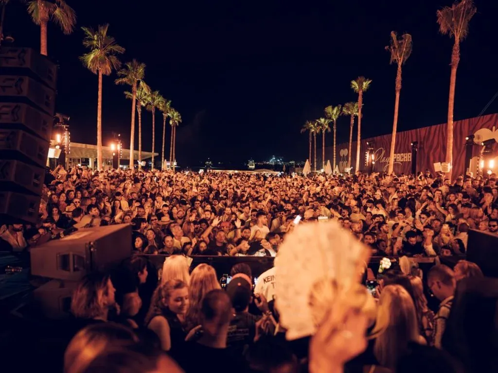 A large crowd of people gathered at a nighttime outdoor event with palm trees in the background, illuminated by bright lights. The scene showcases excitement, with many attendees dancing and holding drinks.