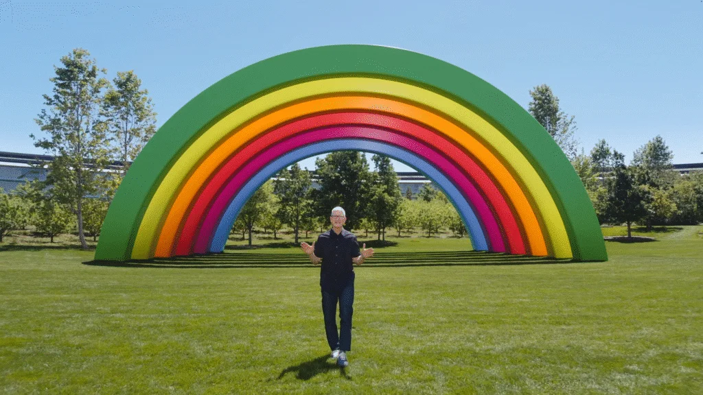 A person stands in front of a large, colorful rainbow sculpture in a park, surrounded by green grass and trees under a clear blue sky.