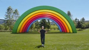 A person stands in front of a large, colorful rainbow sculpture in a park, surrounded by green grass and trees under a clear blue sky.