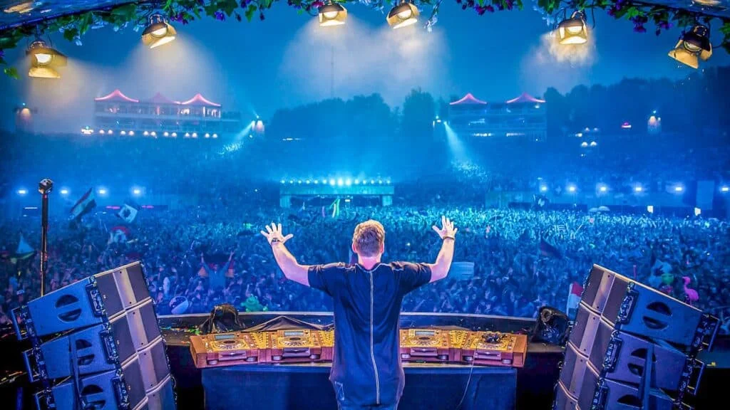 A DJ stands on stage with arms raised, performing in front of a large crowd illuminated by blue lighting and stage lights, with flags waving in the audience and a backdrop of festival structures in the distance.