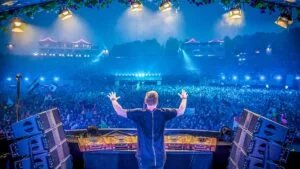 A DJ stands on stage with arms raised, performing in front of a large crowd illuminated by blue lighting and stage lights, with flags waving in the audience and a backdrop of festival structures in the distance.