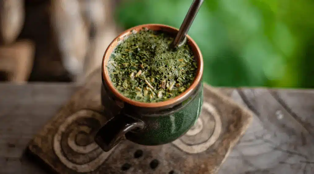 A close-up of a traditional ceramic mug filled with dark green, frothy beverage, resting on a wooden surface with intricate designs.