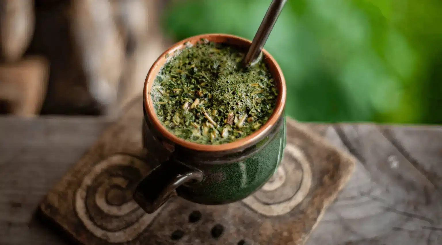 A close-up of a traditional ceramic mug filled with dark green, frothy beverage, resting on a wooden surface with intricate designs.