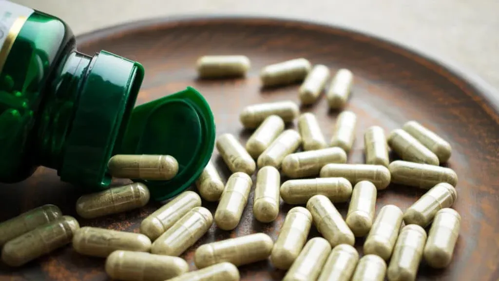 A green bottle of capsules tipped over on a wooden plate, with numerous herbal capsules scattered around.