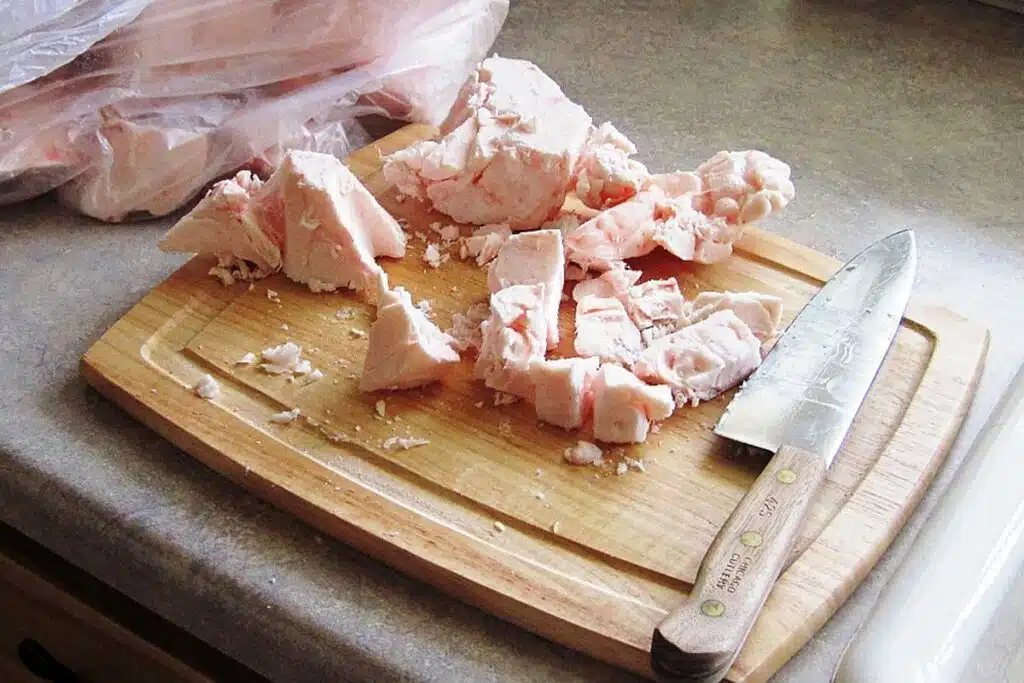 Chunks of raw fat are scattered on a wooden cutting board beside a sharp knife, with a plastic bag of more fat in the background.
