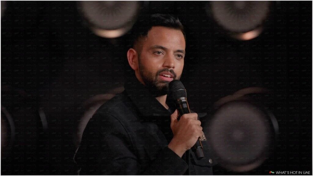 A person with a beard holding a microphone, speaking on stage with a dark background and circular lights.