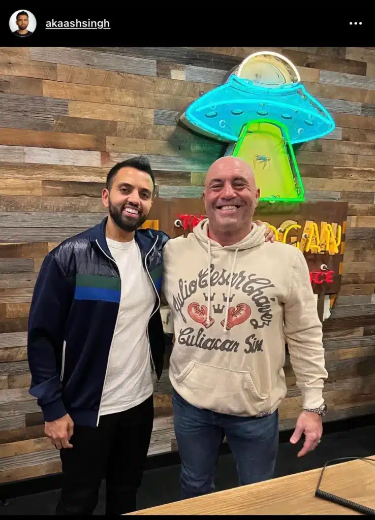 Two men (Akaash Singh on JRE with Joe Rogan) posing together, smiling for the camera in front of a wooden wall with a colorful neon sign of a UFO. One man, wearing a dark jacket, has a beard and short hair, while the other is dressed in a light hoodie and has a shaved head.