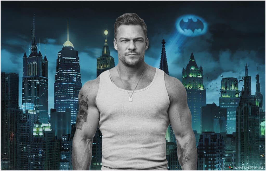 A muscular Alan Ritchson wearing a gray tank top stands in front of a city skyline at night, featuring skyscrapers and the Bat-Signal projected into the sky.