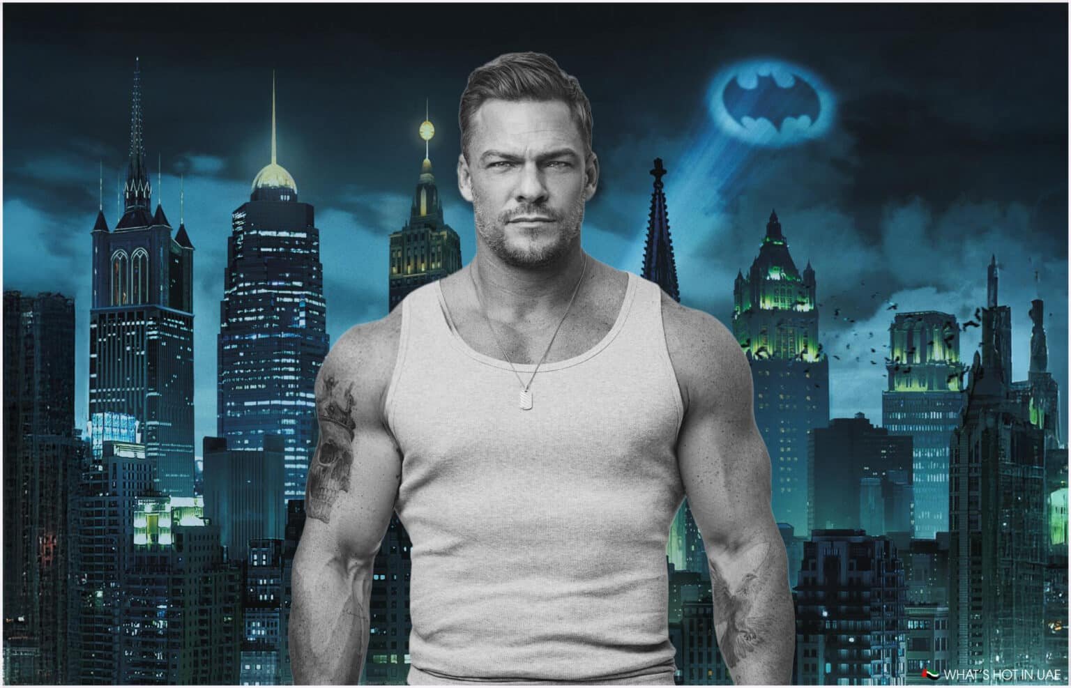 A muscular Alan Ritchson wearing a gray tank top stands in front of a city skyline at night, featuring skyscrapers and the Bat-Signal projected into the sky.