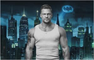 A muscular Alan Ritchson wearing a gray tank top stands in front of a city skyline at night, featuring skyscrapers and the Bat-Signal projected into the sky.