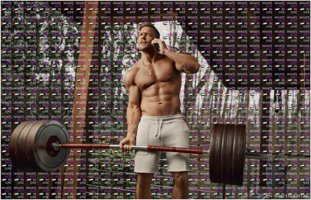 A muscular man is lifting a heavily loaded barbell while talking on a cell phone. He is wearing white shorts and appears to be in a gym setting. The background is overlaid with the text 'WHAT'S HOT IN UAE' repeated multiple times.