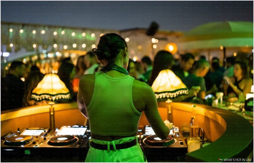 A DJ wearing a white tank top is performing at a lively outdoor venue, surrounded by a crowd of people and illuminated by warm lighting, including a decorative lamp on the DJ booth.